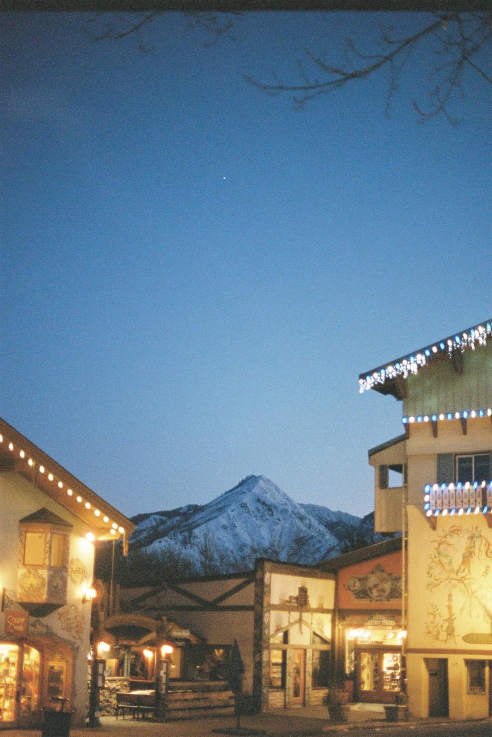 A snow-capped mountain rises behind buildings adorned with string lights under a twilight sky.