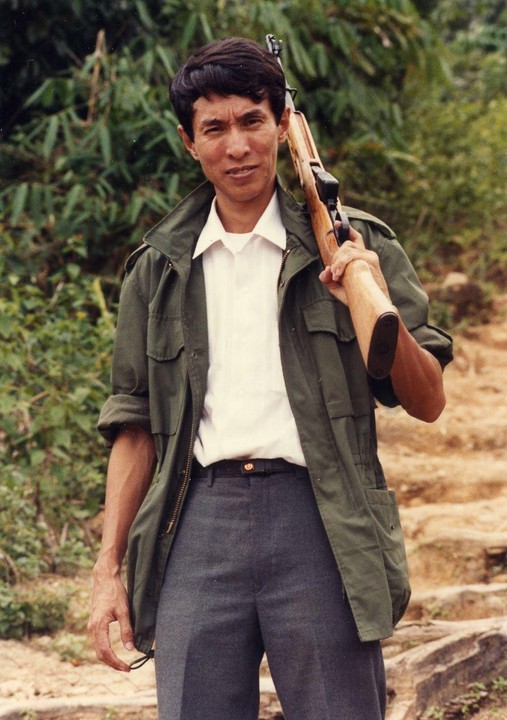 A man in a green jacket and white shirt holds a rifle over his shoulder while standing outdoors.