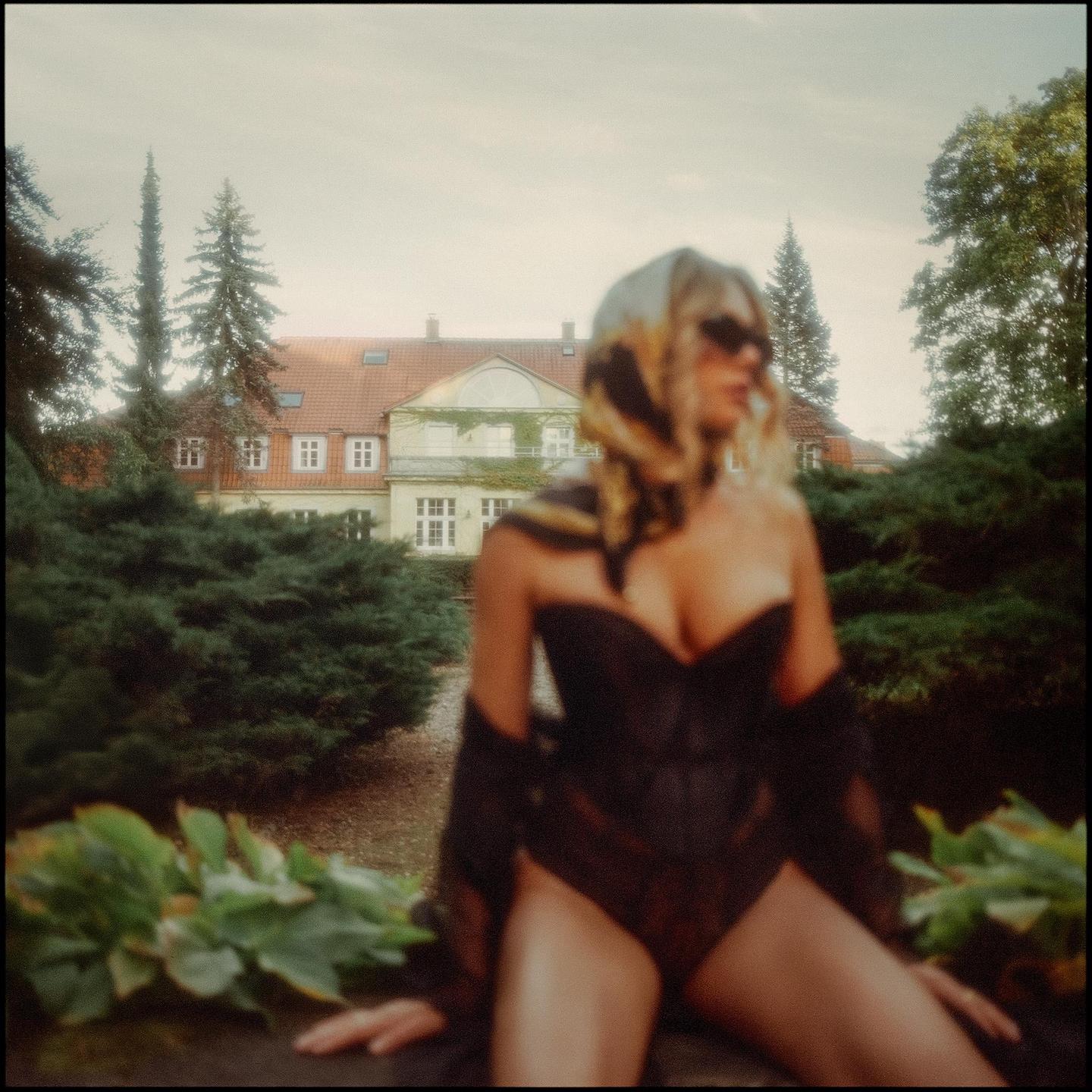 A woman in a black corset and scarf sits in front of a large house with a red roof.