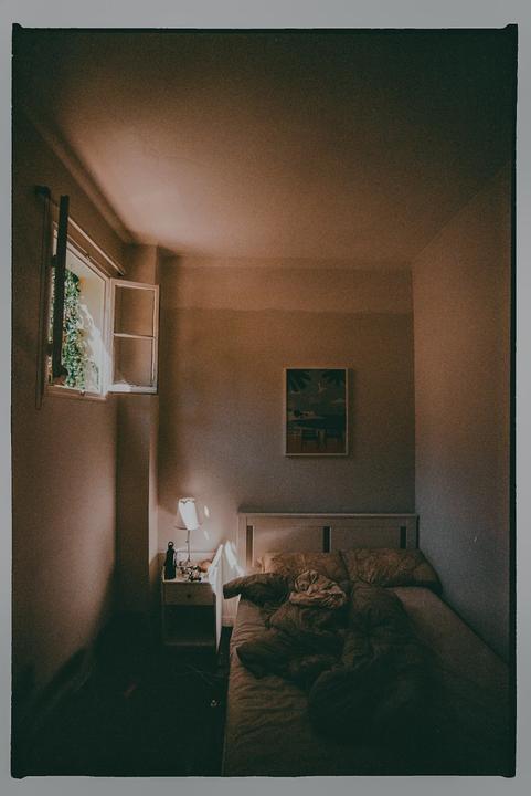 Sunlight streams through an open window into a bedroom with an unmade bed and a bedside table with a lamp.