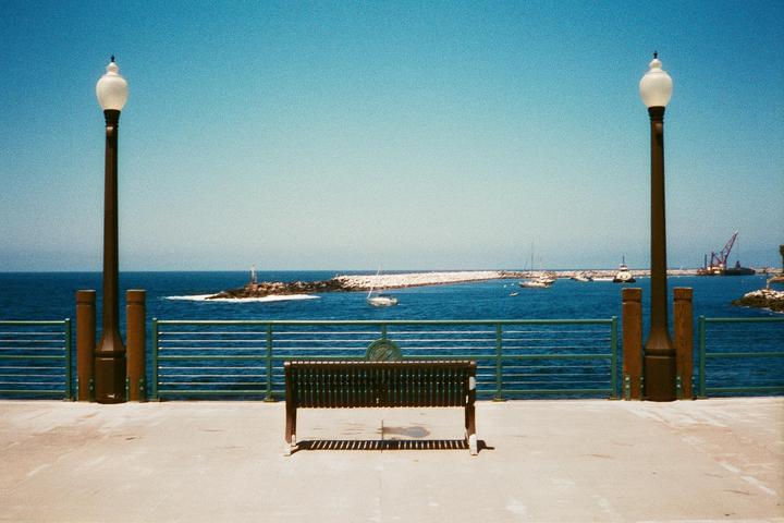 A lone bench sits on a pier overlooking a harbor with boats and a breakwater under a clear sky.