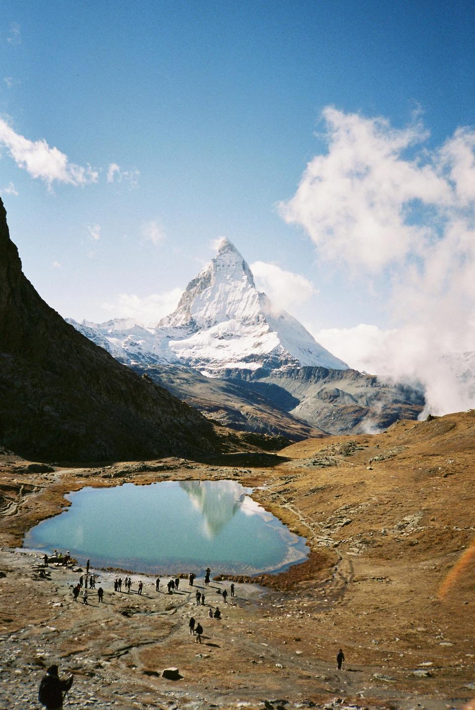 A snow-capped mountain peak is reflected in a small lake surrounded by dry, rocky terrain with many people standing near the water.