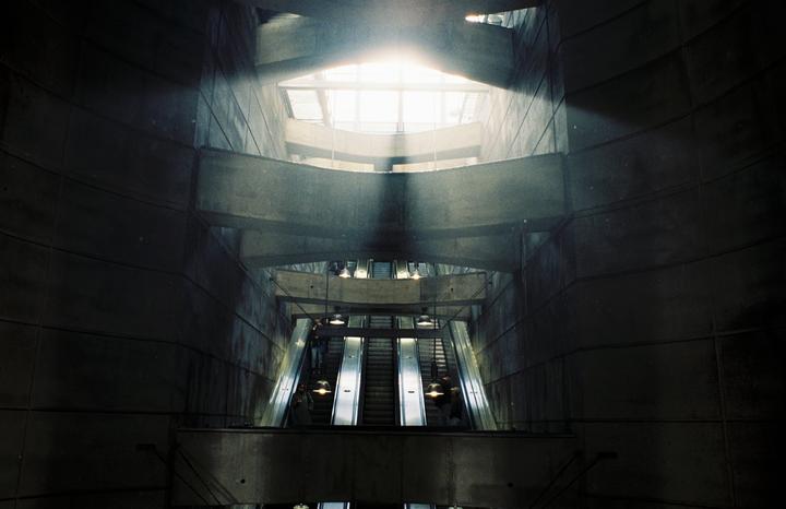 Sunlight streams into a large, dark metro station with multiple escalators and people.