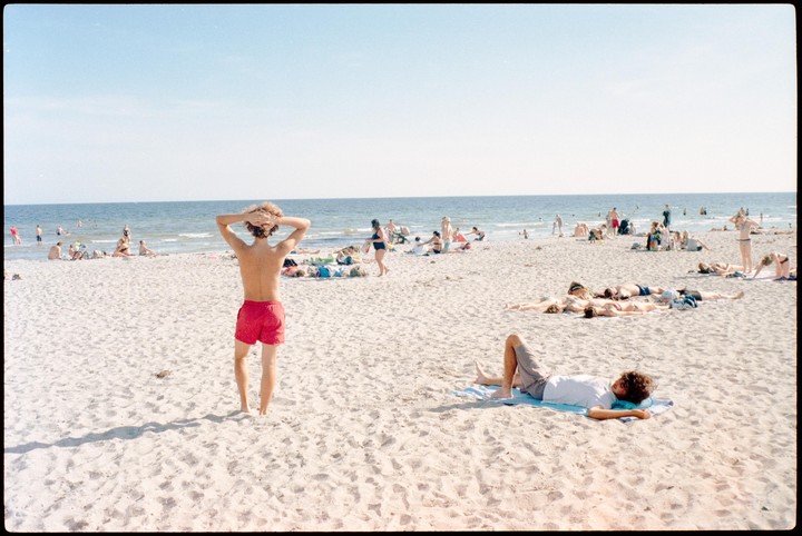 A man in red shorts stands on a sandy beach looking out at the ocean where other people are swimming and sunbathing.