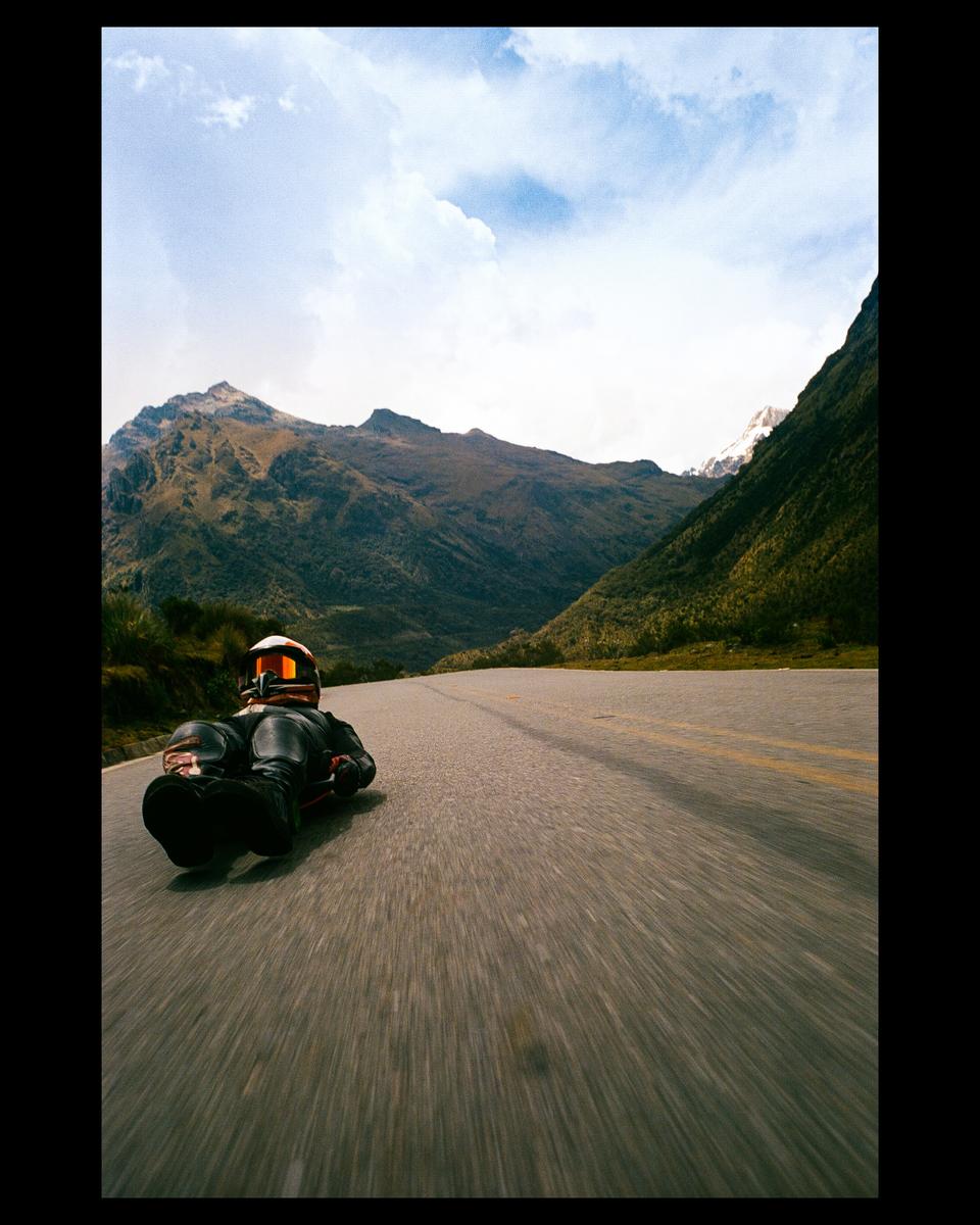 A person in a full-body suit and helmet lies on a street luge on a road with mountains in the background.