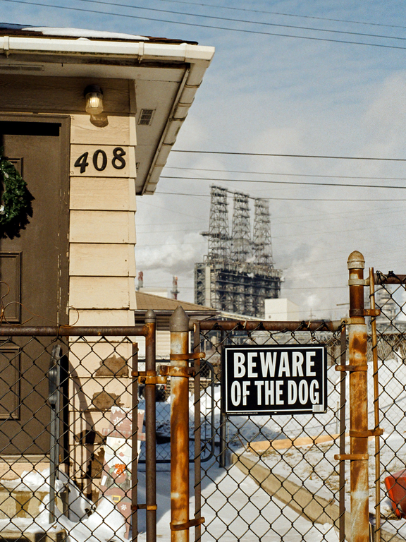 A "Beware of the Dog" sign hangs on a rusty chain-link fence in front of a house with the number 408, with an industrial structure visible in the background.