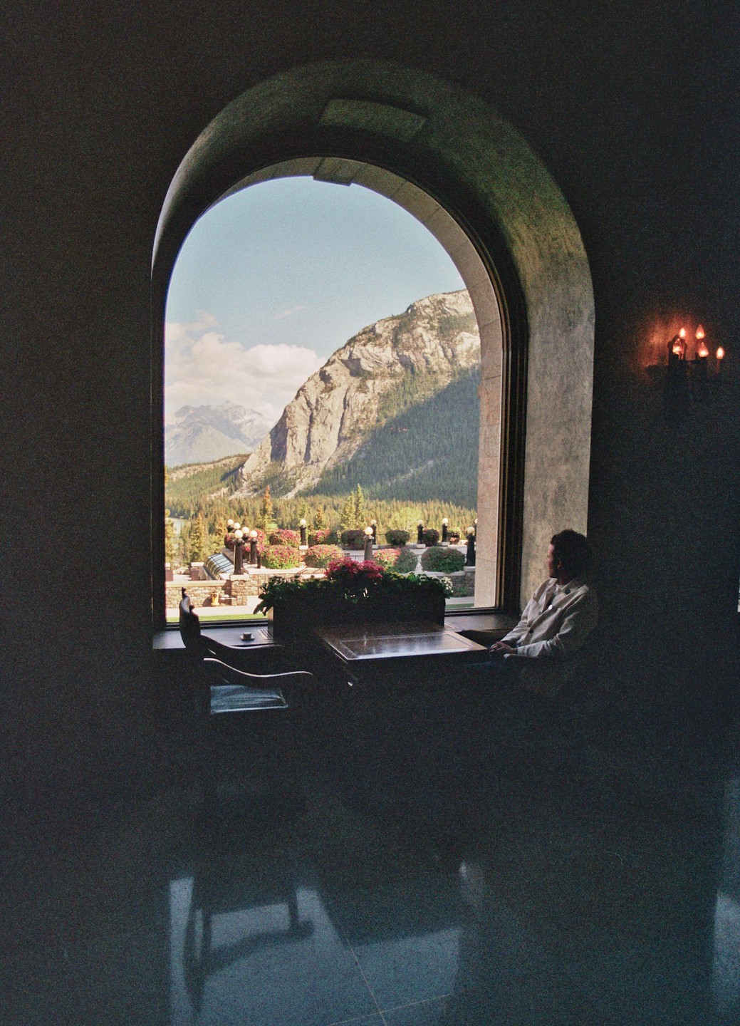 A man sits at a table by a large arched window overlooking a mountain landscape with a garden.