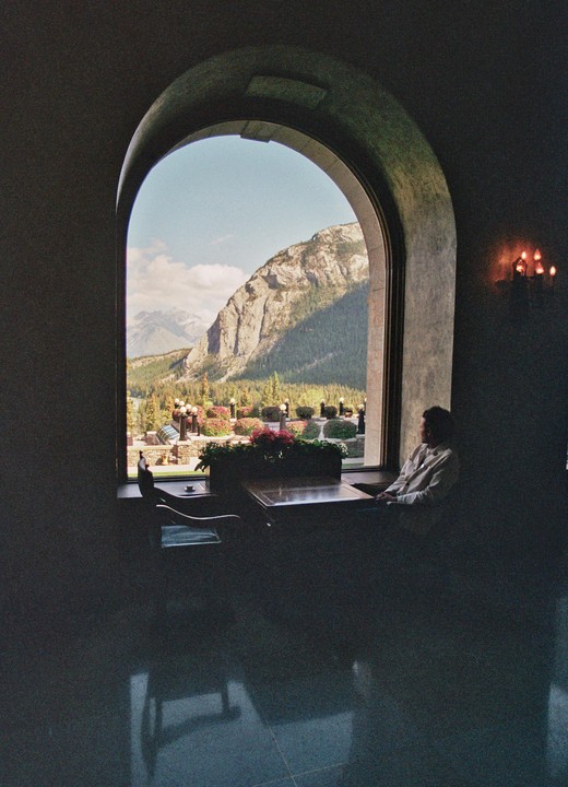 A man sits at a table by a large arched window overlooking a mountain landscape with a garden.