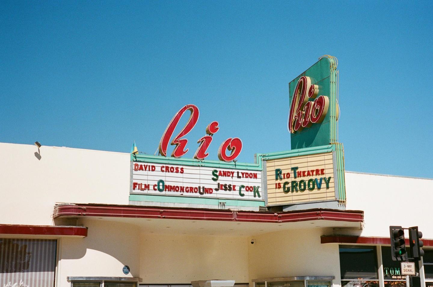 The Rio Theatre marquee with red neon lettering is visible against a clear blue sky.