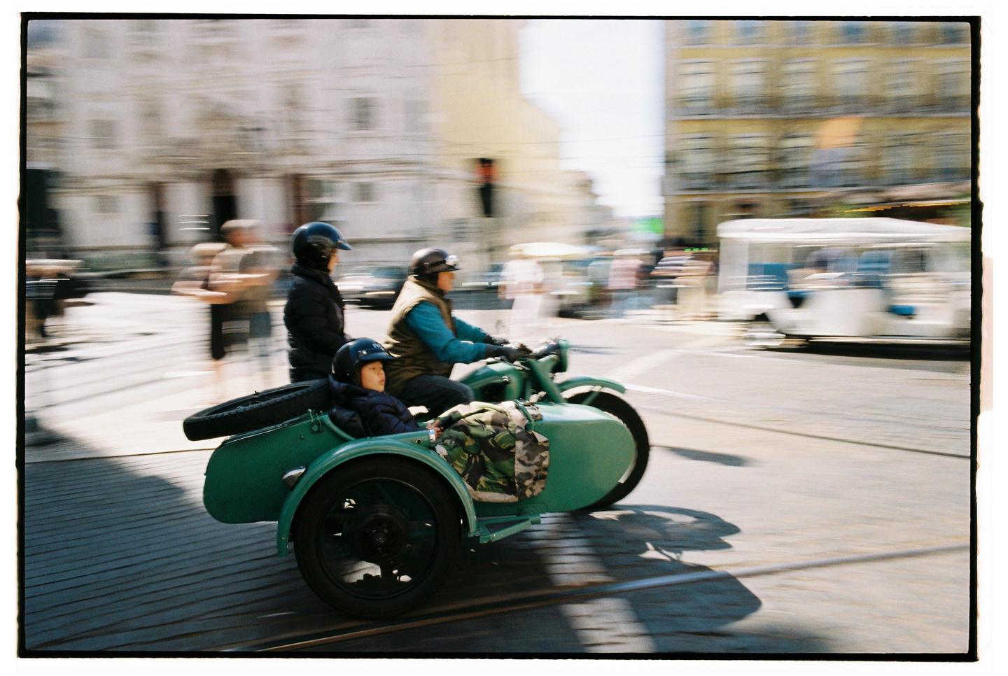 A man drives a green motorcycle with a sidecar carrying a child and another man down a city street.