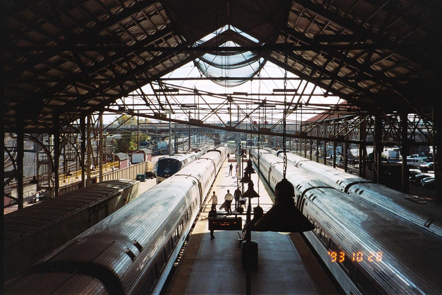 Two trains are parked at a station with people walking on the platform under a large, arched roof.