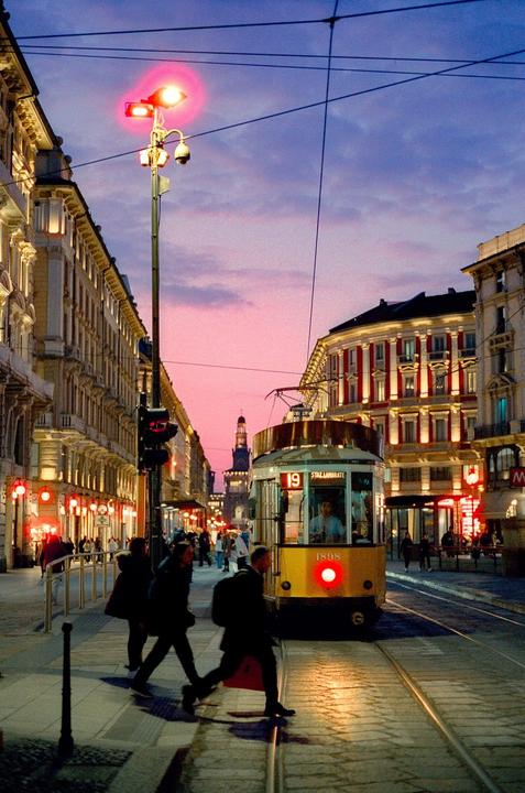 A yellow tram is stopped on a city street with people walking by and buildings lit up under a pink and purple sky.