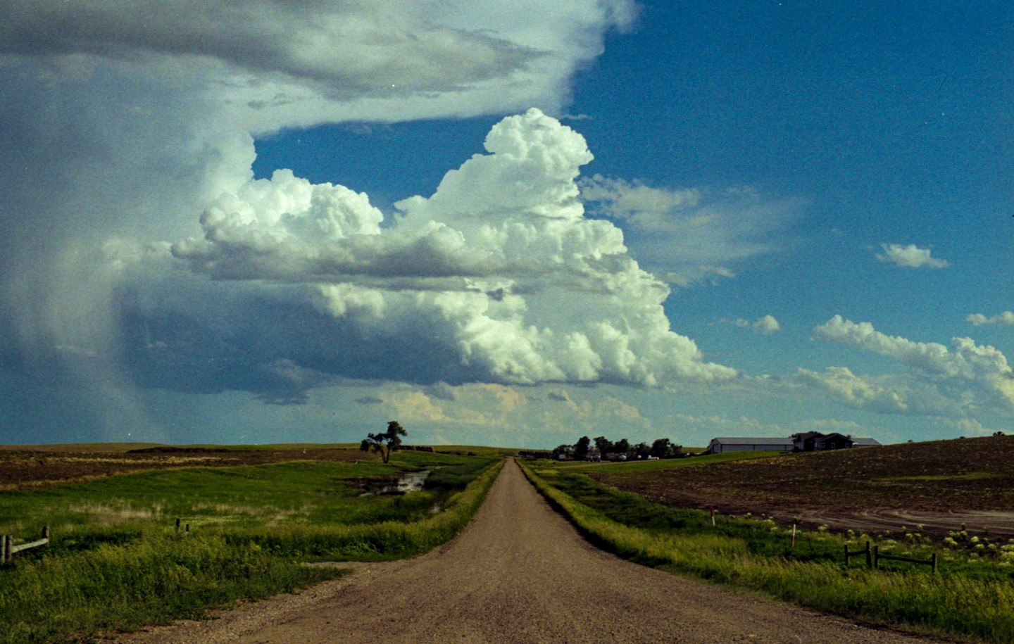 A long dirt road leads through a rural landscape under a dramatic sky with large cumulus clouds.