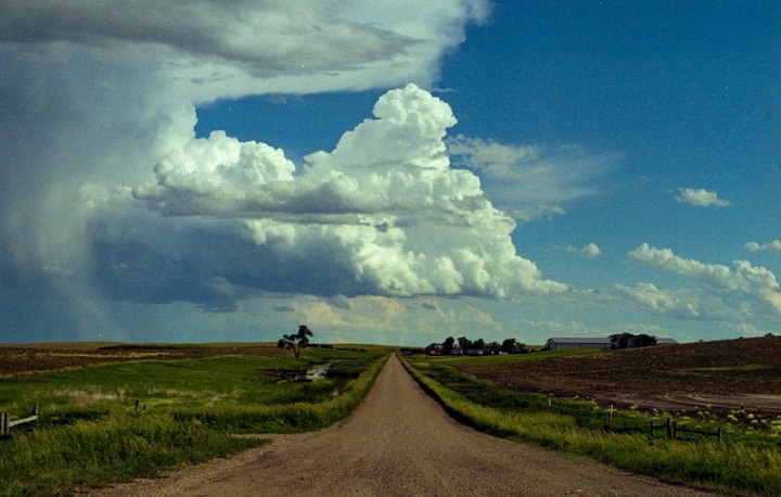 A long dirt road leads through a rural landscape under a dramatic sky with large cumulus clouds.