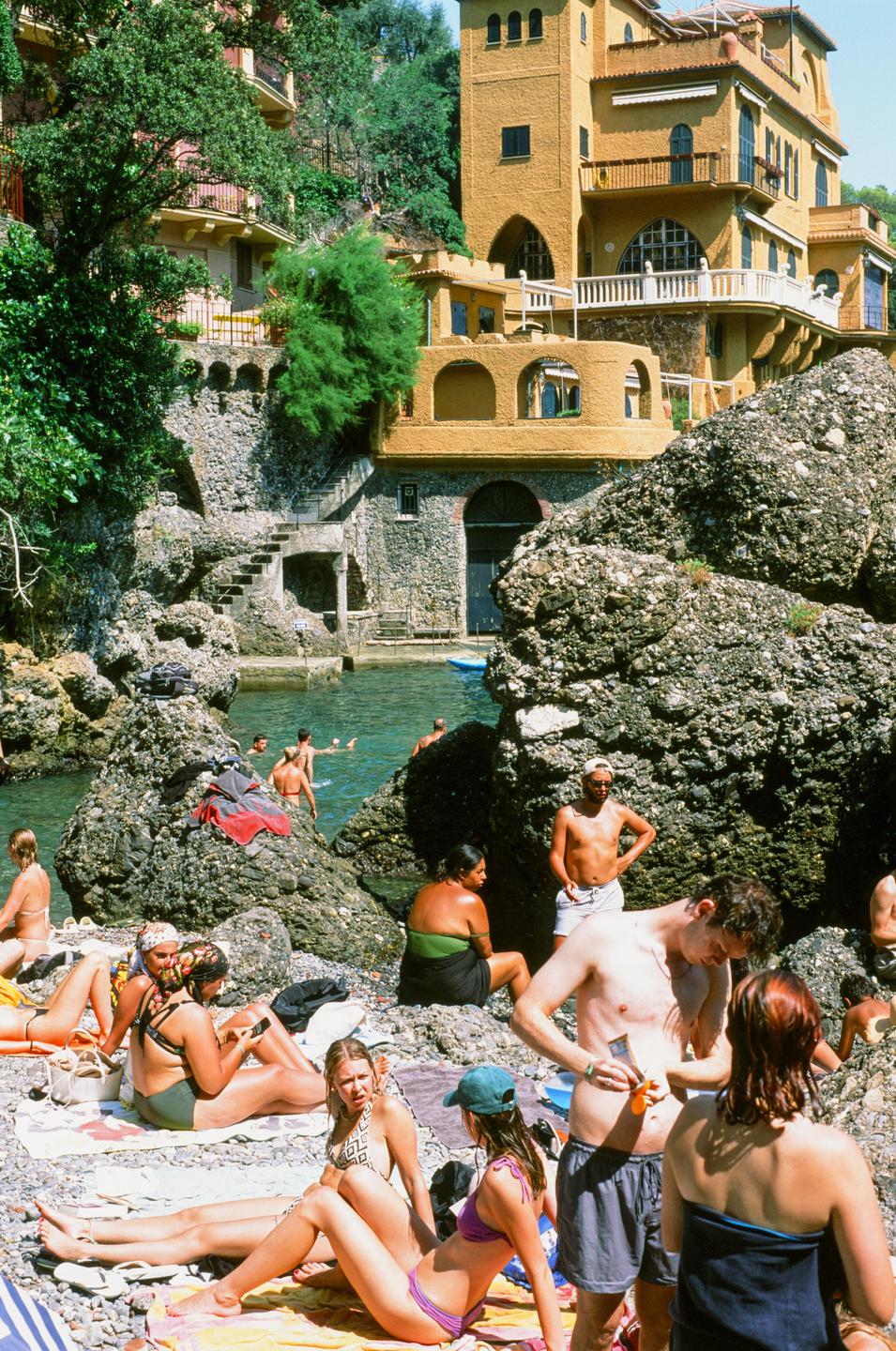 A group of people relax on a rocky beach next to a body of water with buildings on a hillside in the background.