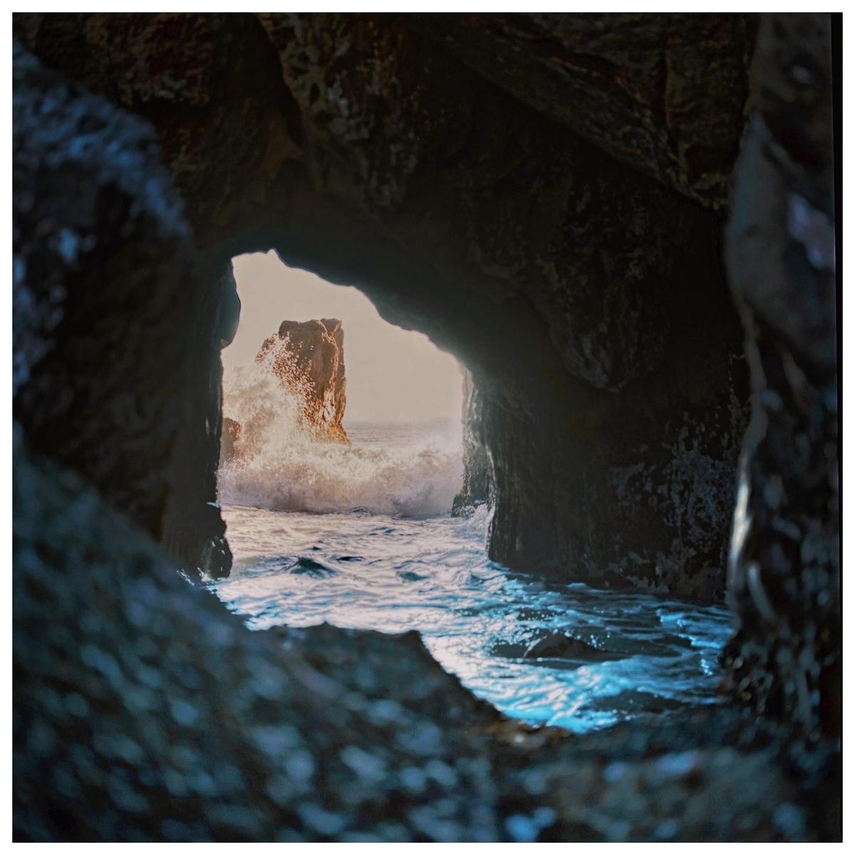 A large wave crashes against a rock formation seen through a dark cave opening.