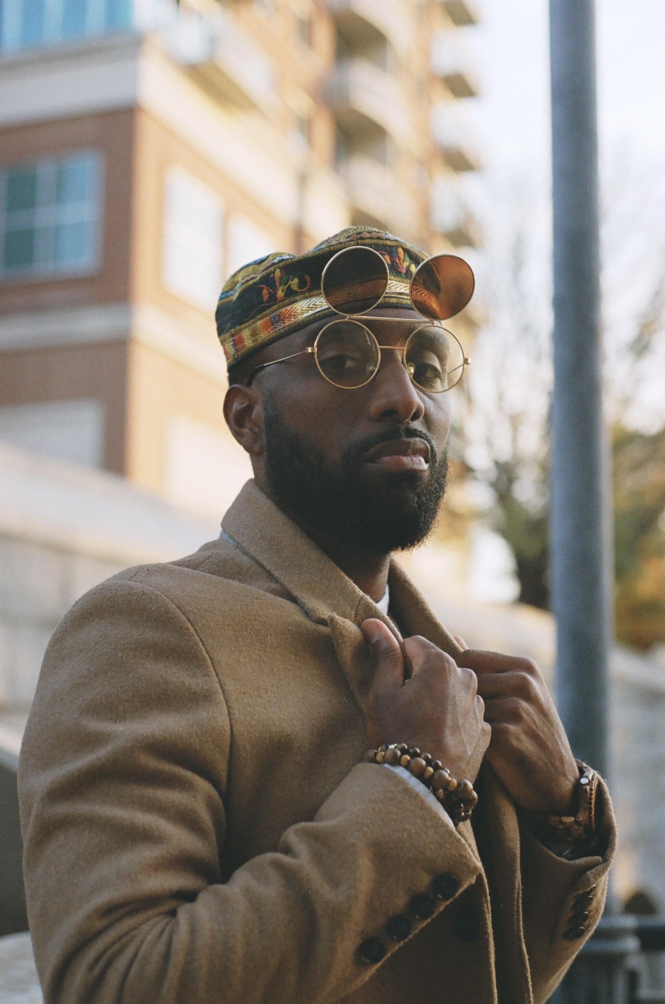 A man wearing a patterned hat, round sunglasses, and a tan coat poses outdoors.