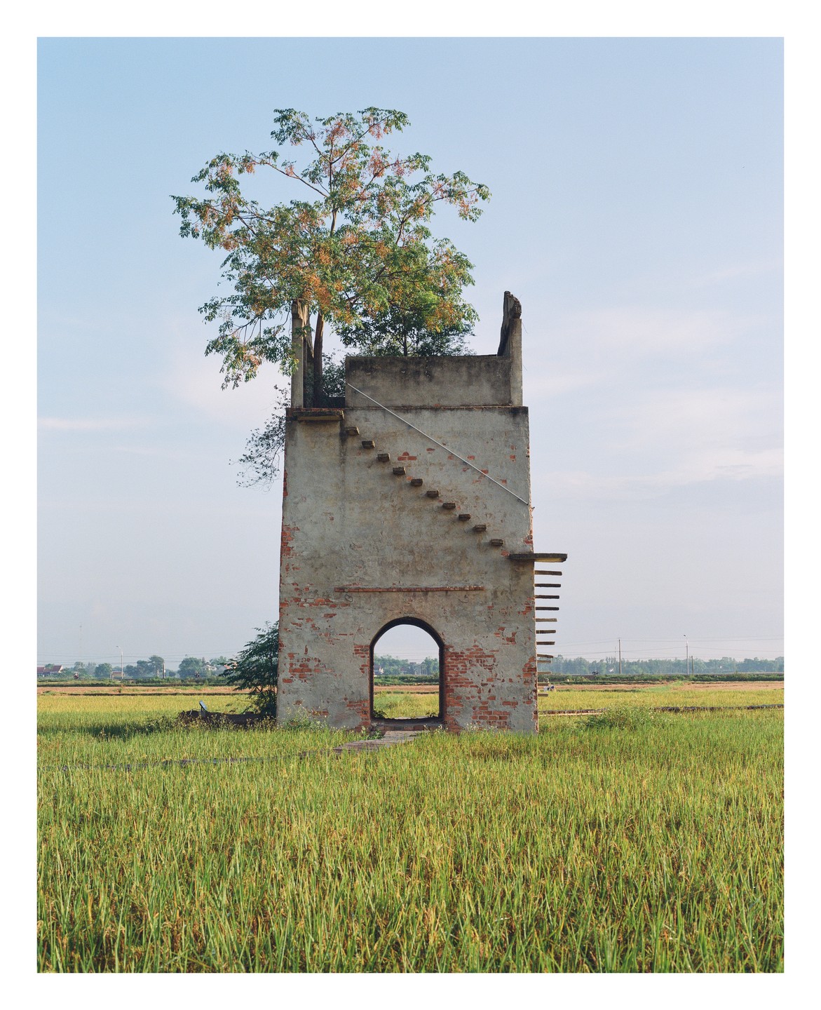 A tall, narrow, abandoned brick structure with an arched doorway and an exterior staircase stands in a field of tall grass under a clear sky.