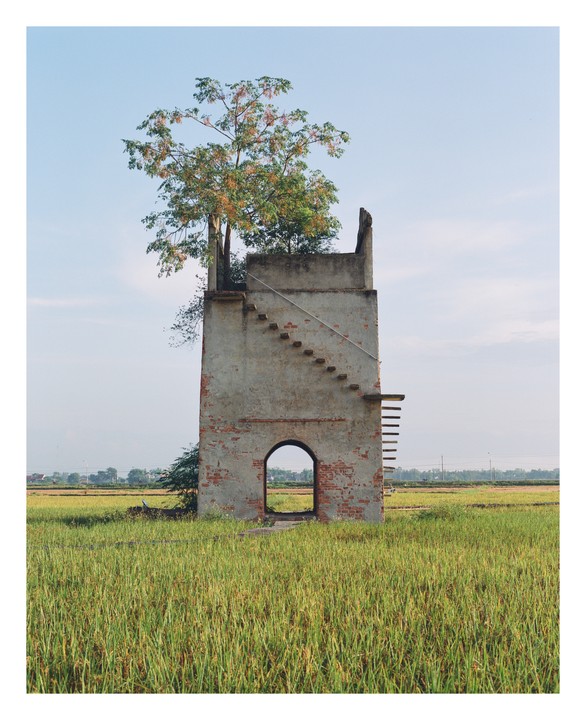 A tall, narrow, abandoned brick structure with an arched doorway and an exterior staircase stands in a field of tall grass under a clear sky.