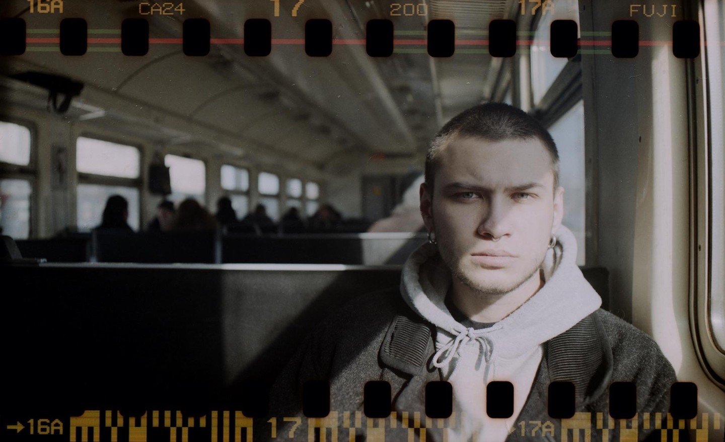 A man with a buzz cut, nose ring, and earrings sits on a train, looking directly at the viewer.