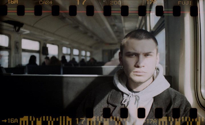 A man with a buzz cut, nose ring, and earrings sits on a train, looking directly at the viewer.