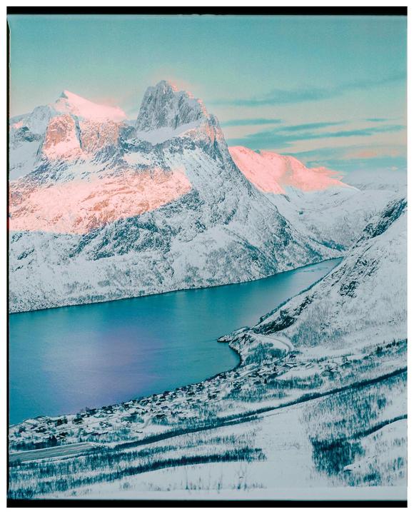 A snow-covered mountain range with a body of water and a small village at its base is visible under a blue sky.