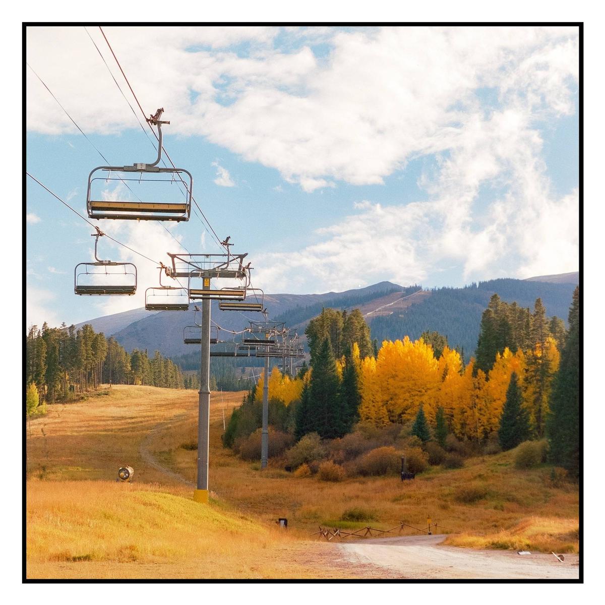 A ski lift ascends a mountain with autumn foliage and a dirt road below.