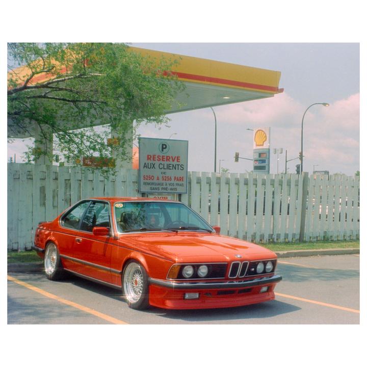 A red car is parked in a lot next to a white fence and a gas station.