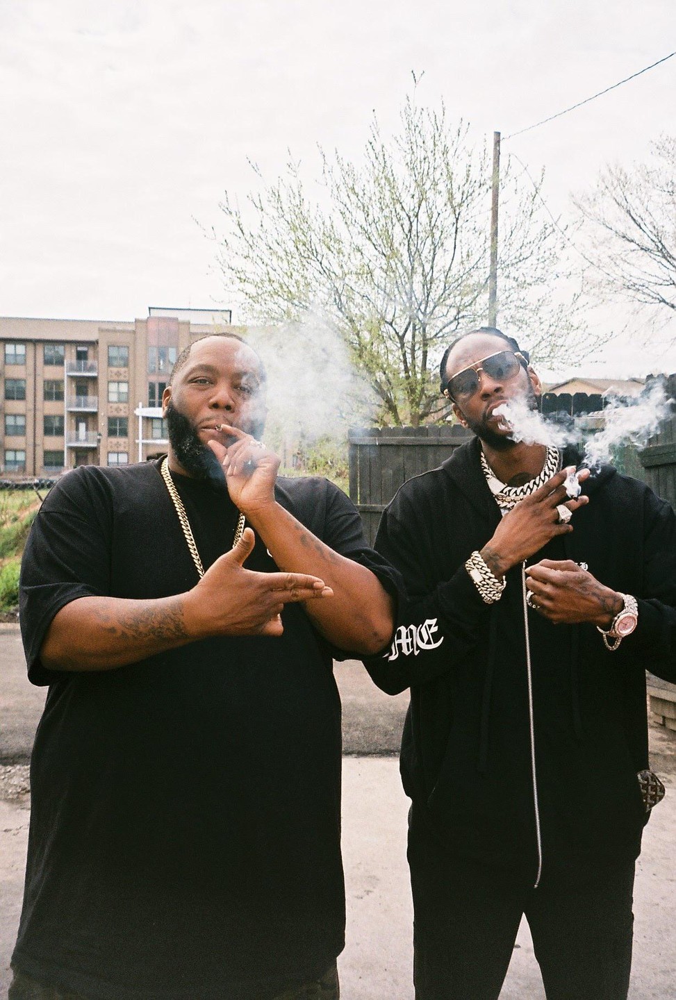 Two men stand outside smoking, with buildings and trees in the background.