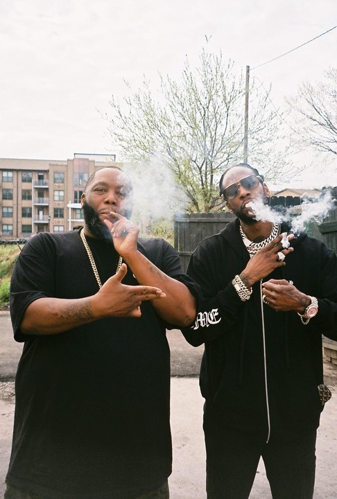 Two men stand outside smoking, with buildings and trees in the background.