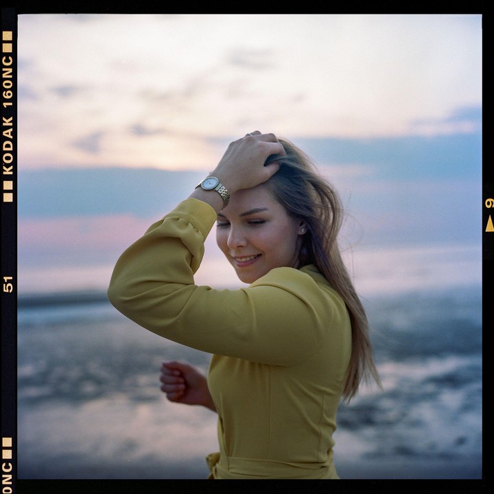 A woman in a yellow dress smiles with her hand in her hair against a soft blue and pink sky.