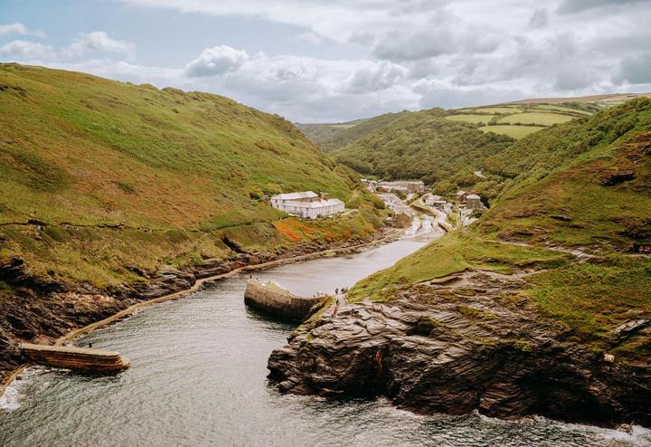 A small village with white buildings is nestled in a valley between green hills, with a river flowing through it and out to the sea.
