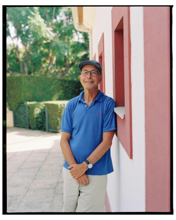 A man wearing a blue polo shirt, a cap, and glasses is standing next to a building with red and white stripes.