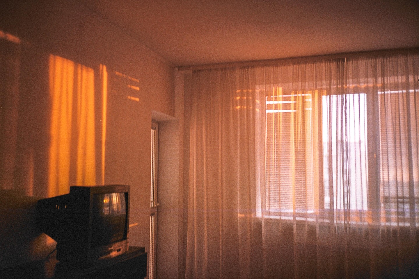 Sunlight streams through a window with sheer curtains and blinds, casting warm light on a television set in a room.