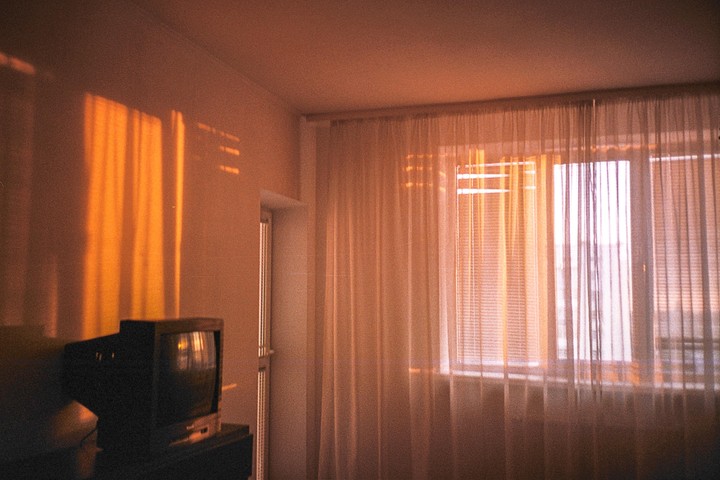 Sunlight streams through a window with sheer curtains and blinds, casting warm light on a television set in a room.