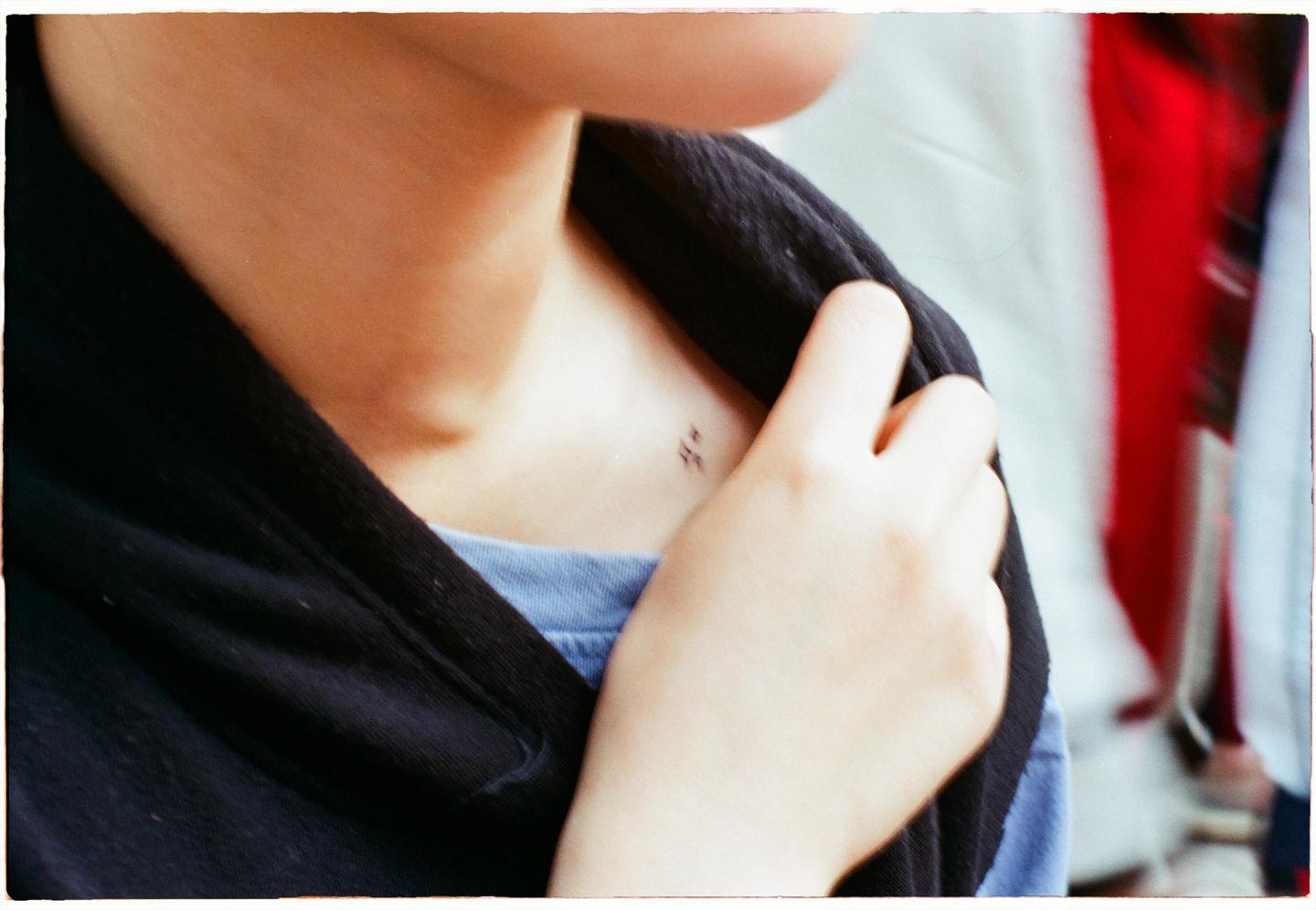 A person's hand rests on their shoulder, revealing a small tattoo on their collarbone.