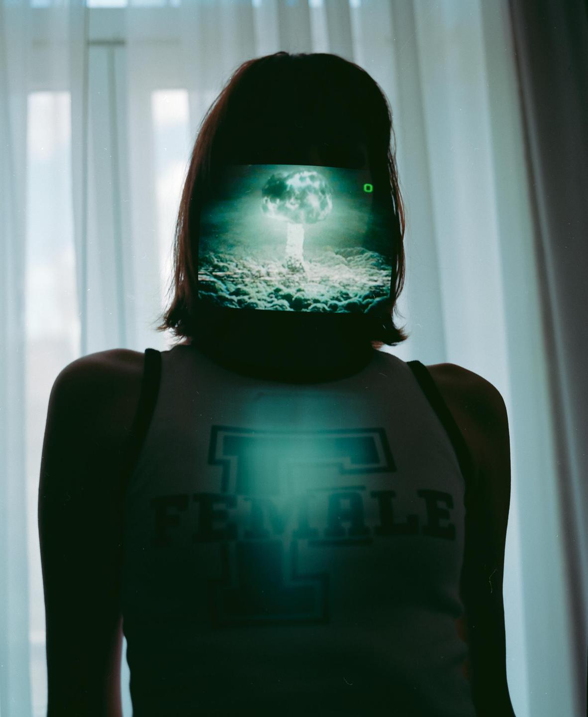 A woman stands with a nuclear explosion projected onto her face and a glowing chest.