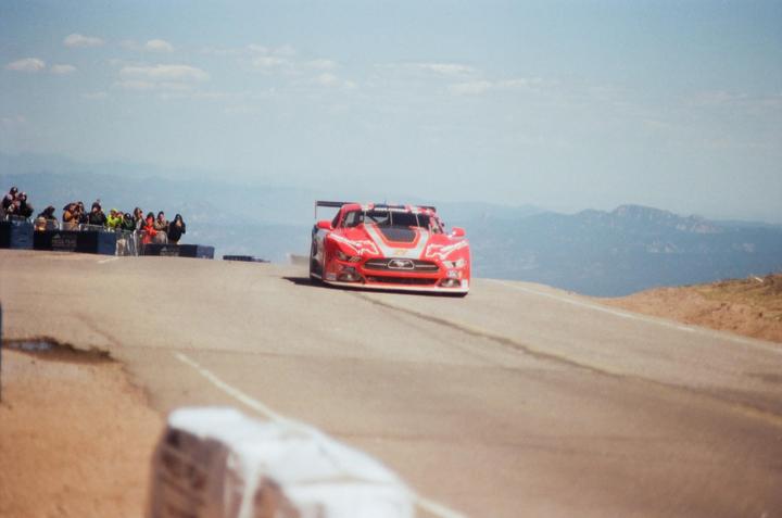 A red race car speeds along a mountain road with spectators and mountains in the background.