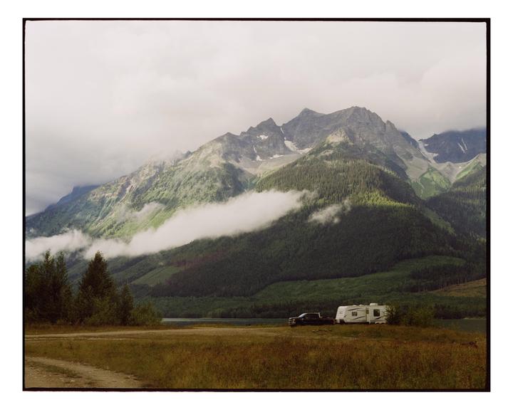 A truck and a camper are parked in a field with a lake and mountains in the background.