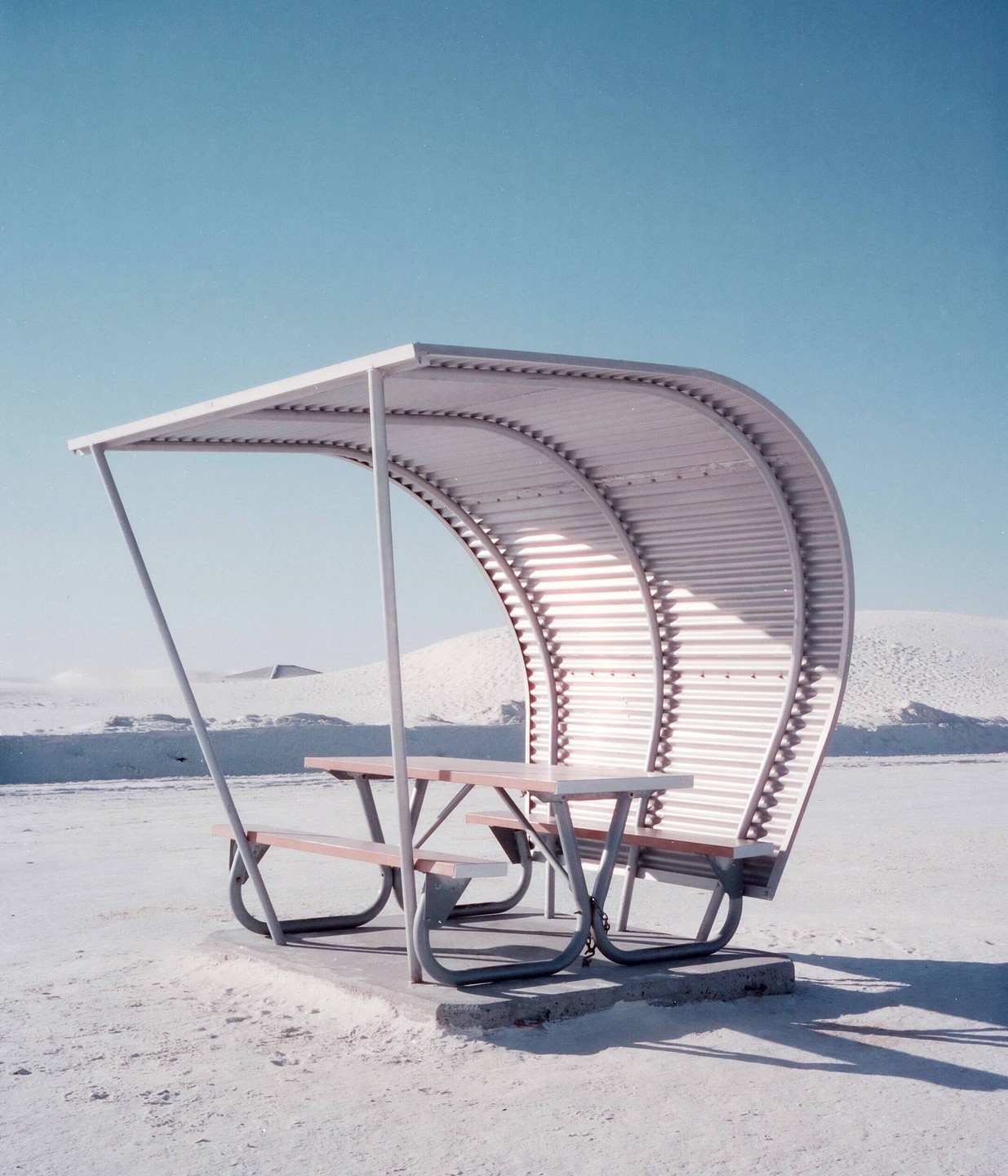 A picnic table with an arched metal roof stands on a white sandy ground under a clear blue sky.