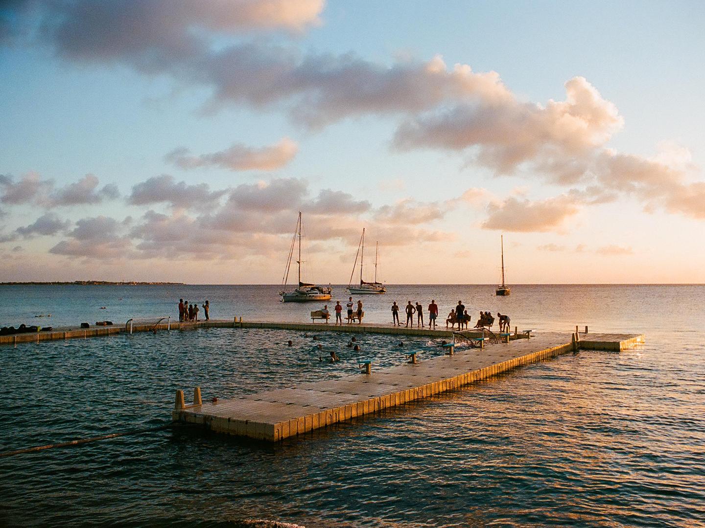 People are playing water polo in a sea pool at sunset, with sailboats visible in the distance.