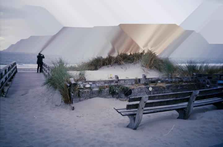 A person stands on a wooden boardwalk looking out at the ocean with sand dunes and a bench in the foreground.