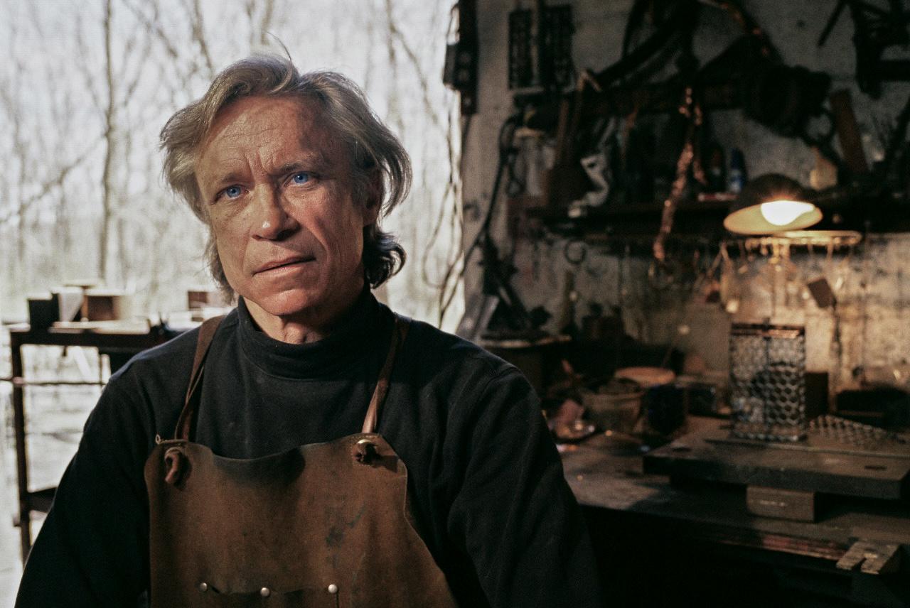 A man wearing an apron looks directly at the viewer in a workshop.