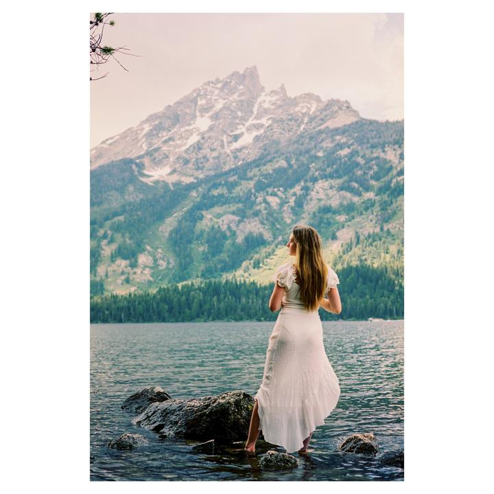 A woman stands in a lake with mountains and a forest in the background.