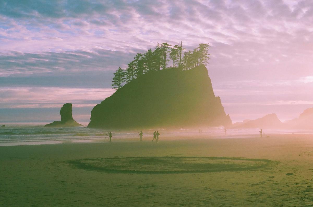 A large rock formation with trees on top stands on a beach with people walking near the water under a colorful sky.