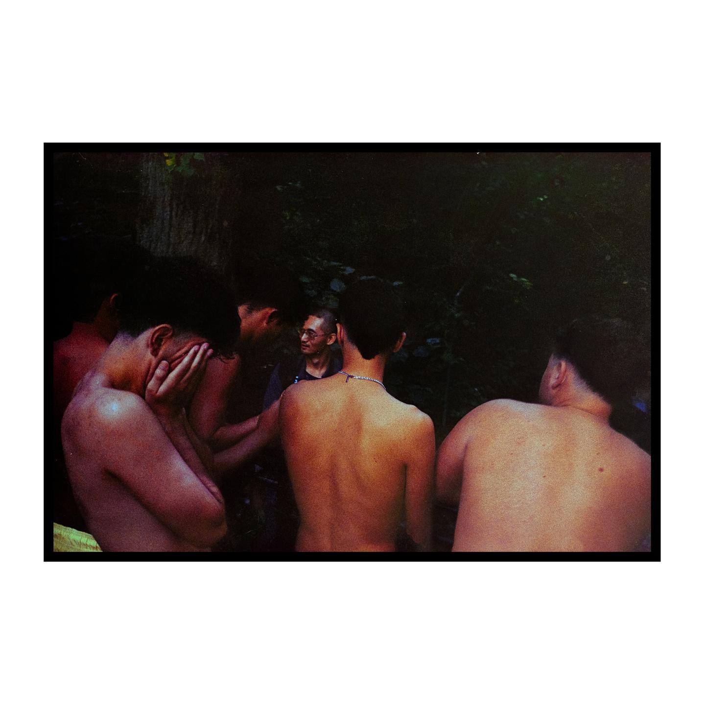 A group of shirtless men are gathered in a dark, wooded area, with one man in the center wearing glasses and a shirt.