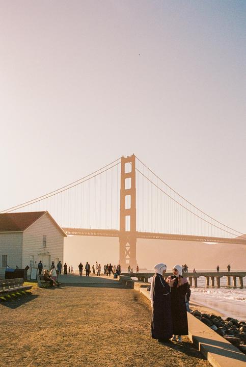 Two women stand on a beach with a bridge and a building in the background.