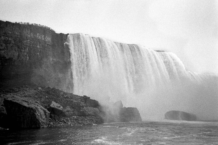 A wide waterfall cascades into a river, with a cliff and a crowd of people visible on the left.