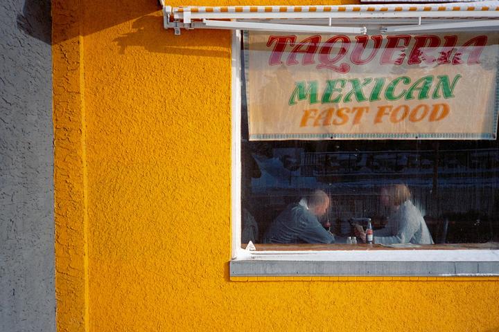 Two people sit inside a taqueria with a sign for Mexican fast food visible in the window.