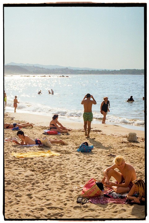 People are relaxing on a sunny beach with sparkling water and distant hills.
